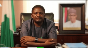 Femi Adesina sitting and staring into the camera, with Buhari's picture at the background